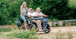 Eine ehrenamtliche Rikscha-Pilotin fährt zwei Seniorinnen für das Projekt „Radeln ohne Alter“ der Bürgerstiftung Braunschweig durch einen Park.